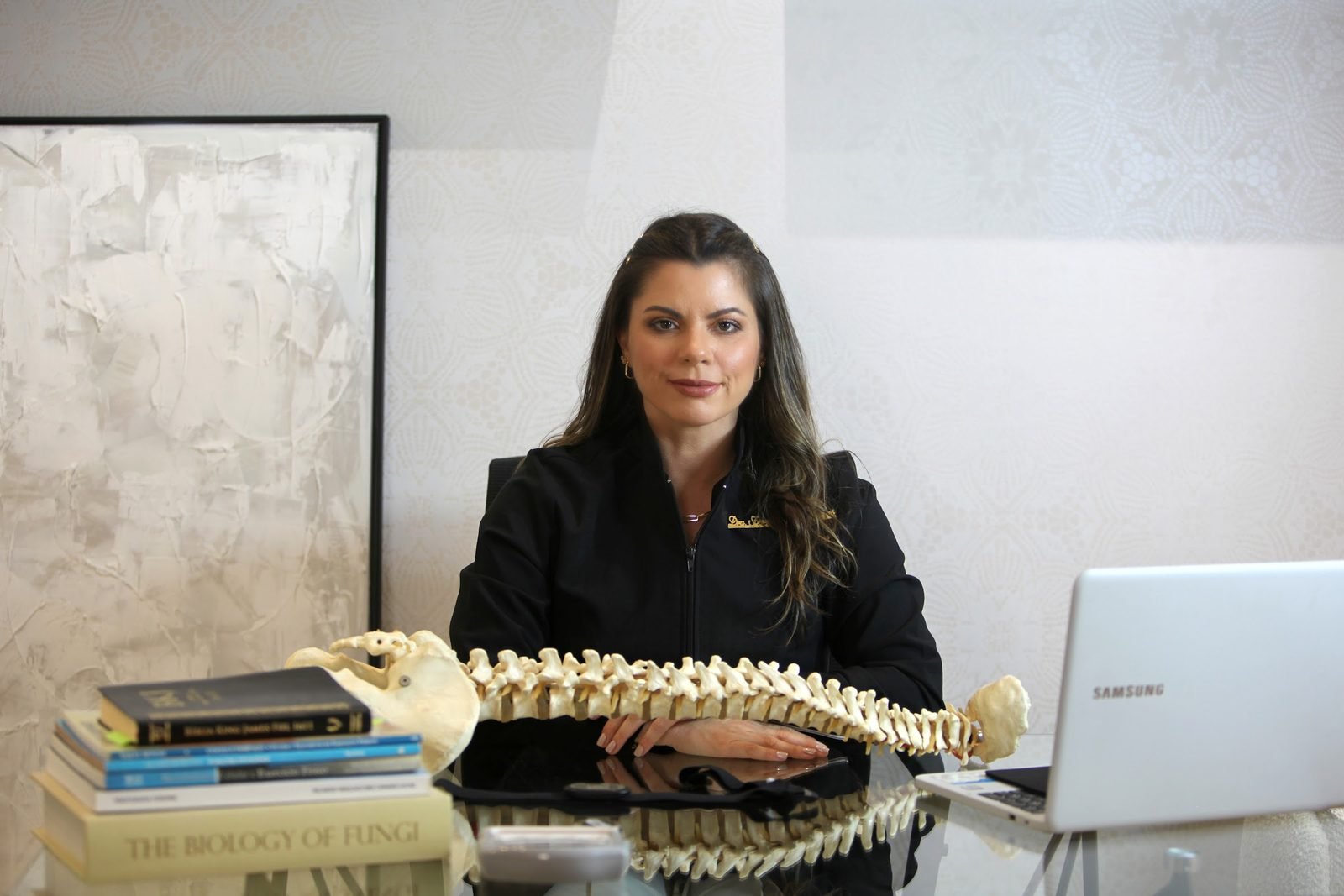 Dra. Fernanda Marques, de jaleco preto, à mesa com um modelo da coluna vertebral, livros e notebook