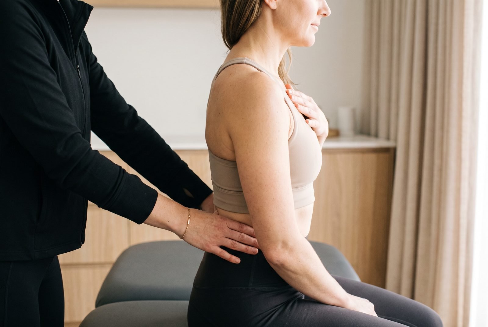 Fisioterapeuta de jaleco preto posiciona as mãos na lombar de uma mulher sentada na maca para corrigir o alinhamento da coluna