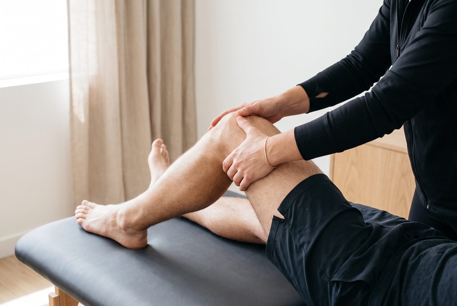 Mãos da fisioterapeuta avaliando o joelho dobrado de um homem deitado na maca